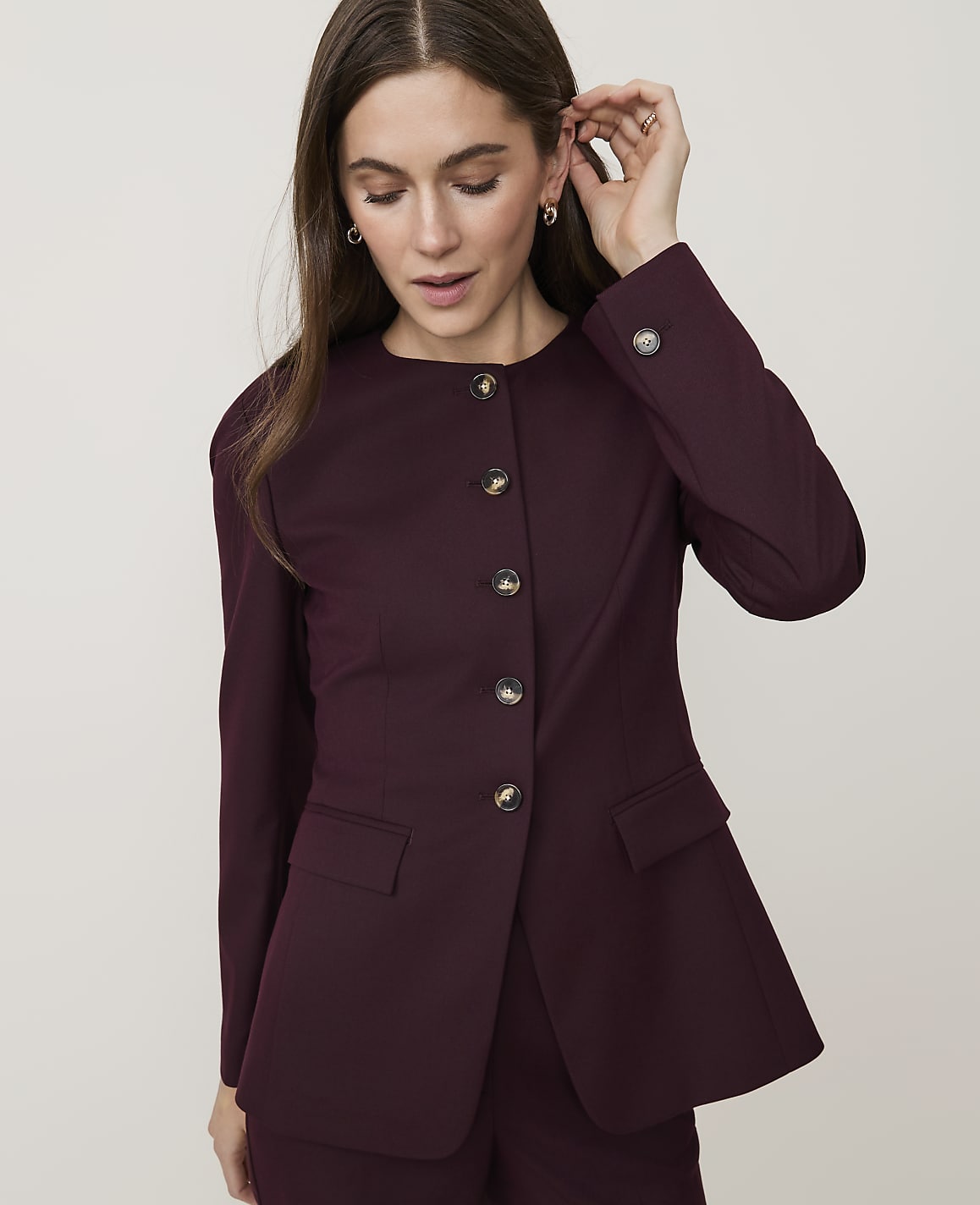 A woman in a burgundy blazer with round buttons adjusts her earring against a plain white background.