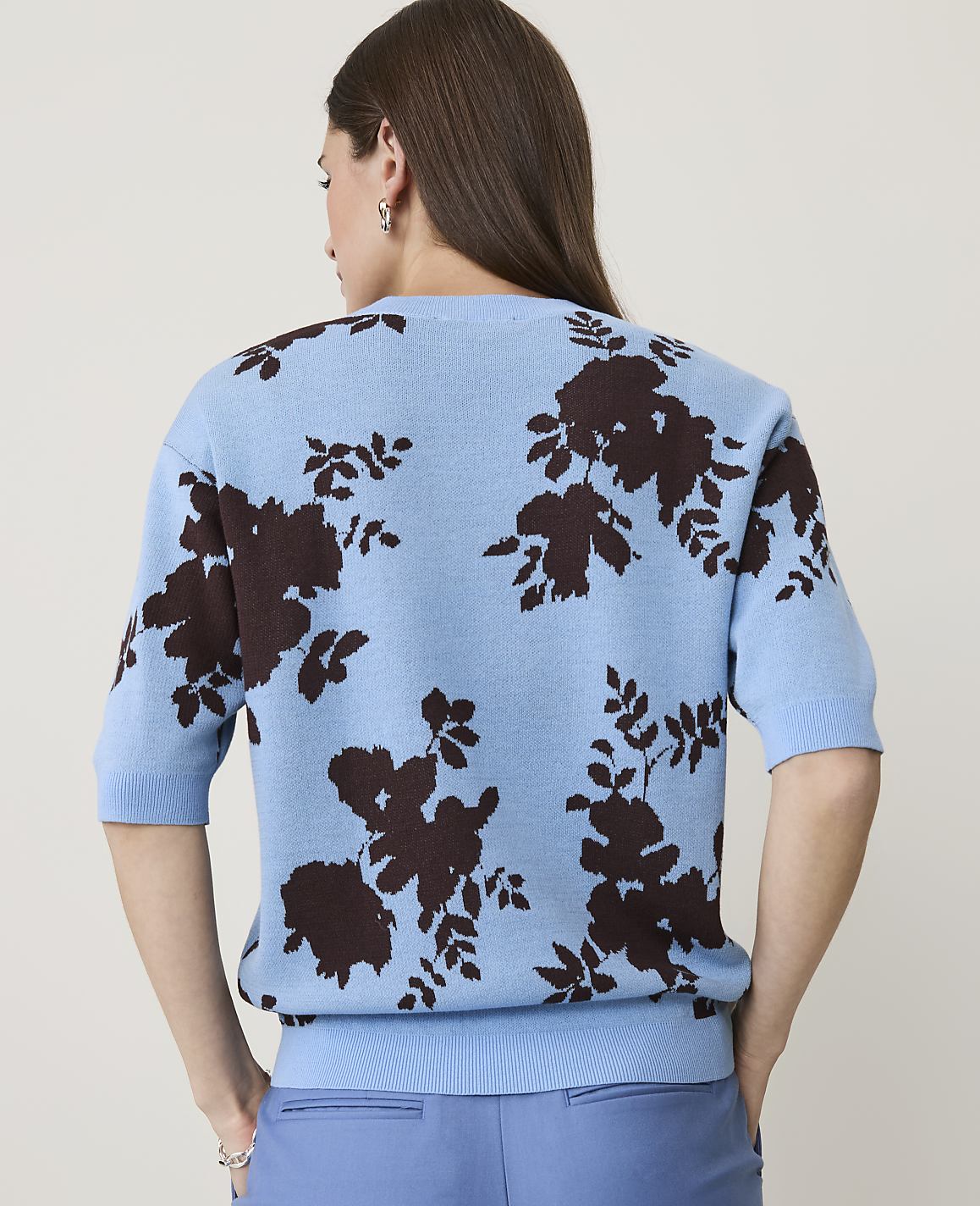 A woman in a light blue floral sweater and blue pants stands against a plain, light background.