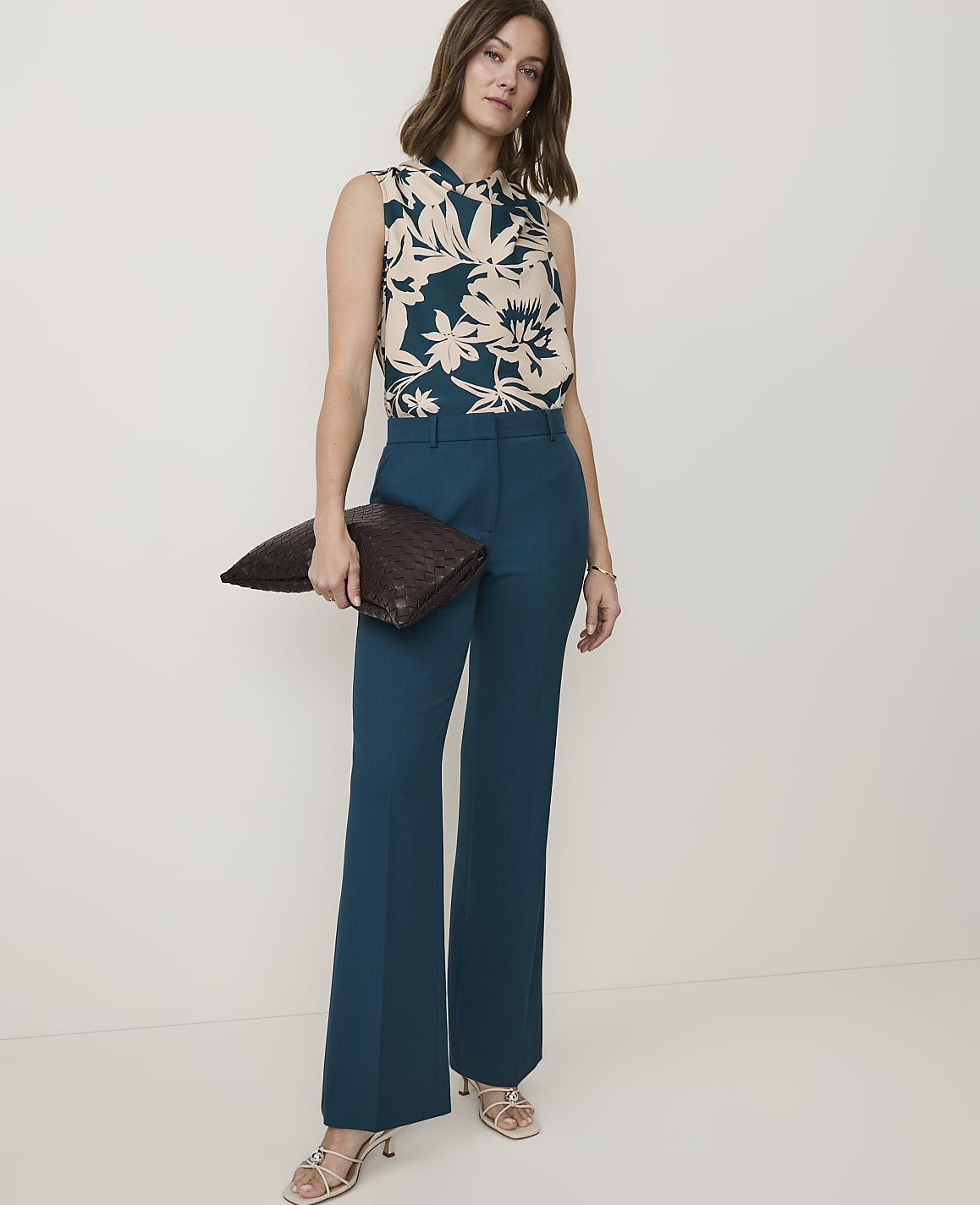 A woman in a floral sleeveless top and teal pants holds a dark clutch in front of a plain background.