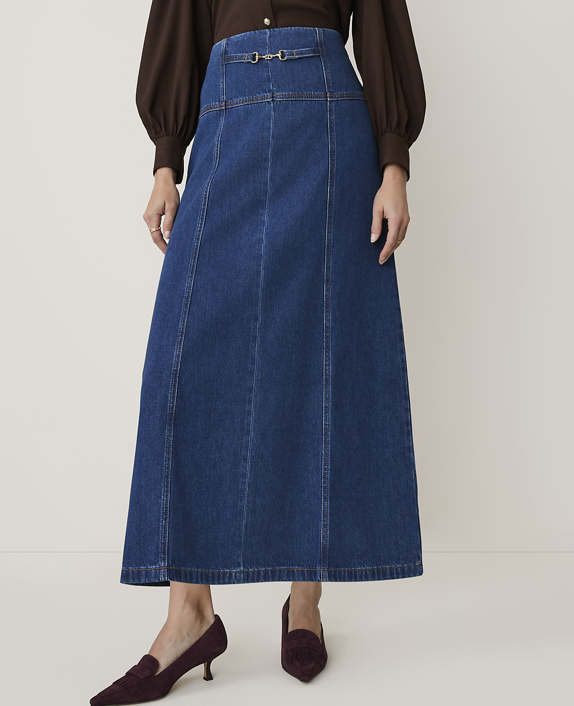 A woman in a brown blouse and a dark denim maxi skirt with a buckle detail stands on a light-colored floor, wearing burgundy loafers.