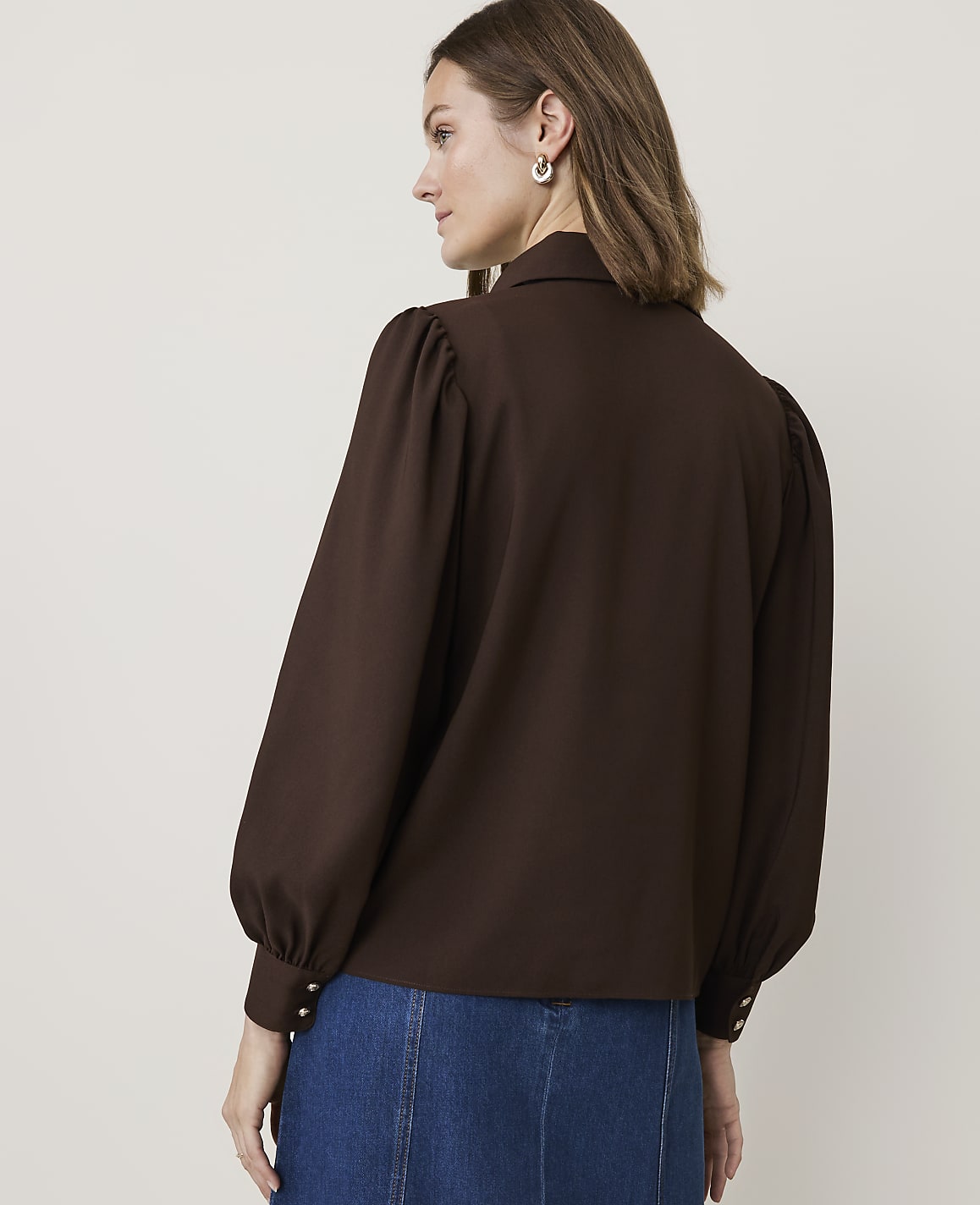 A woman in a brown puff-sleeved blouse and denim skirt stands against a plain beige background.