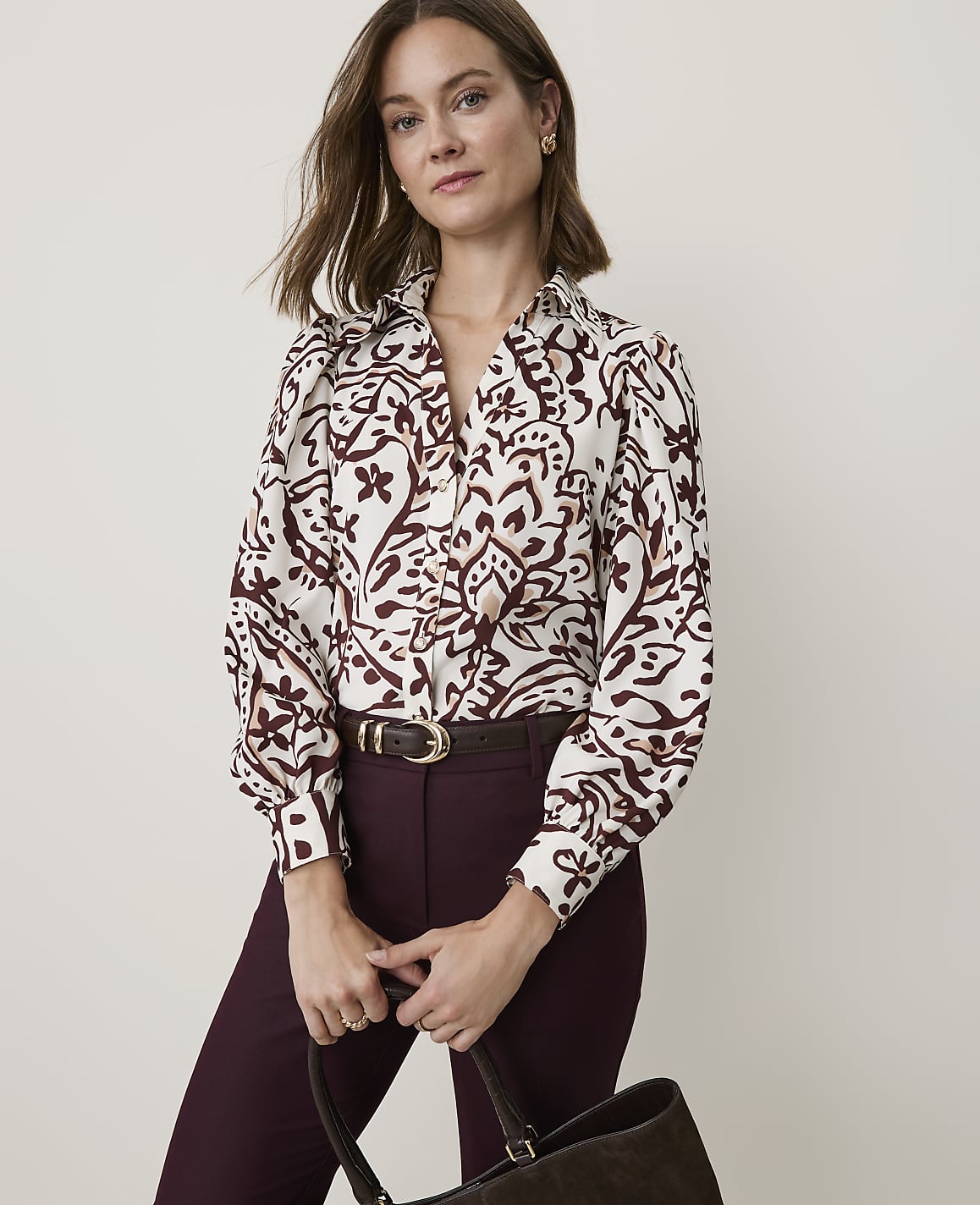 A woman in a patterned blouse and burgundy pants holds a dark brown bag in front of a plain, light-colored background.