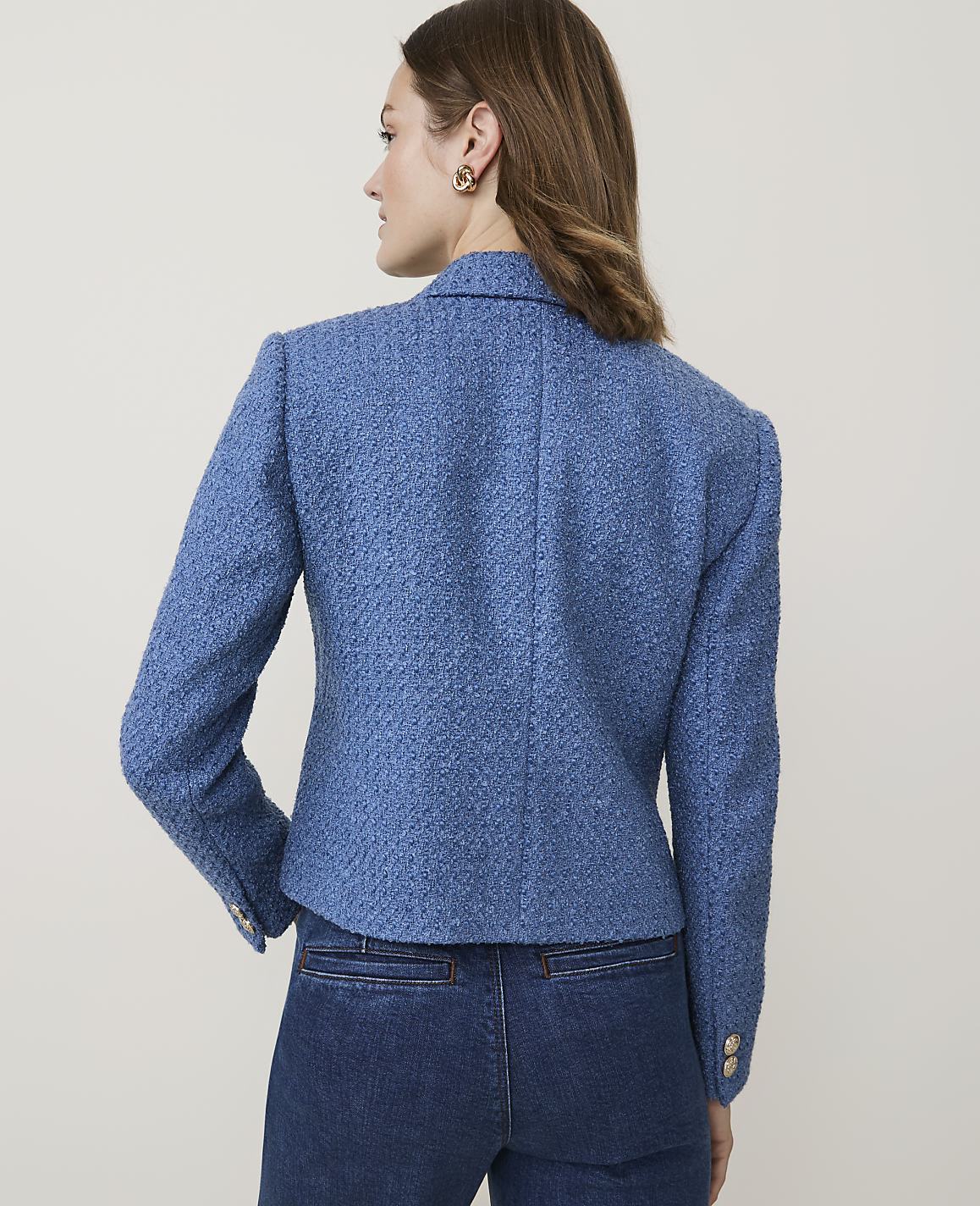 A woman in a blue tweed blazer and denim jeans stands against a plain, light-colored background.