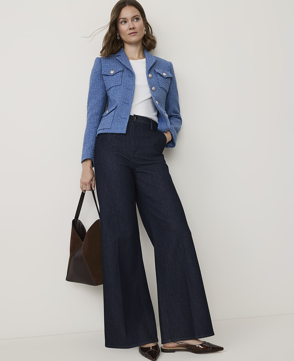 A woman in a blue tweed jacket and dark wide-leg pants stands against a plain white background, holding a brown tote bag.