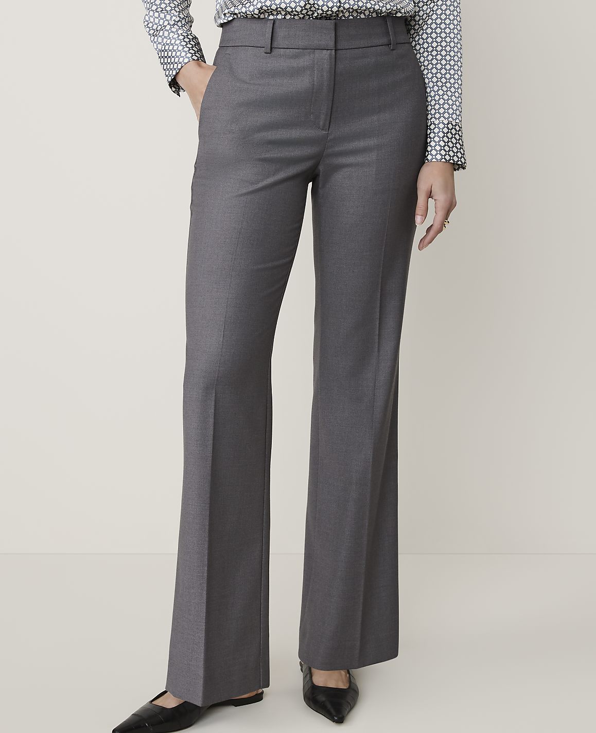 A person stands in gray dress pants and a patterned shirt against a plain, light-colored background.