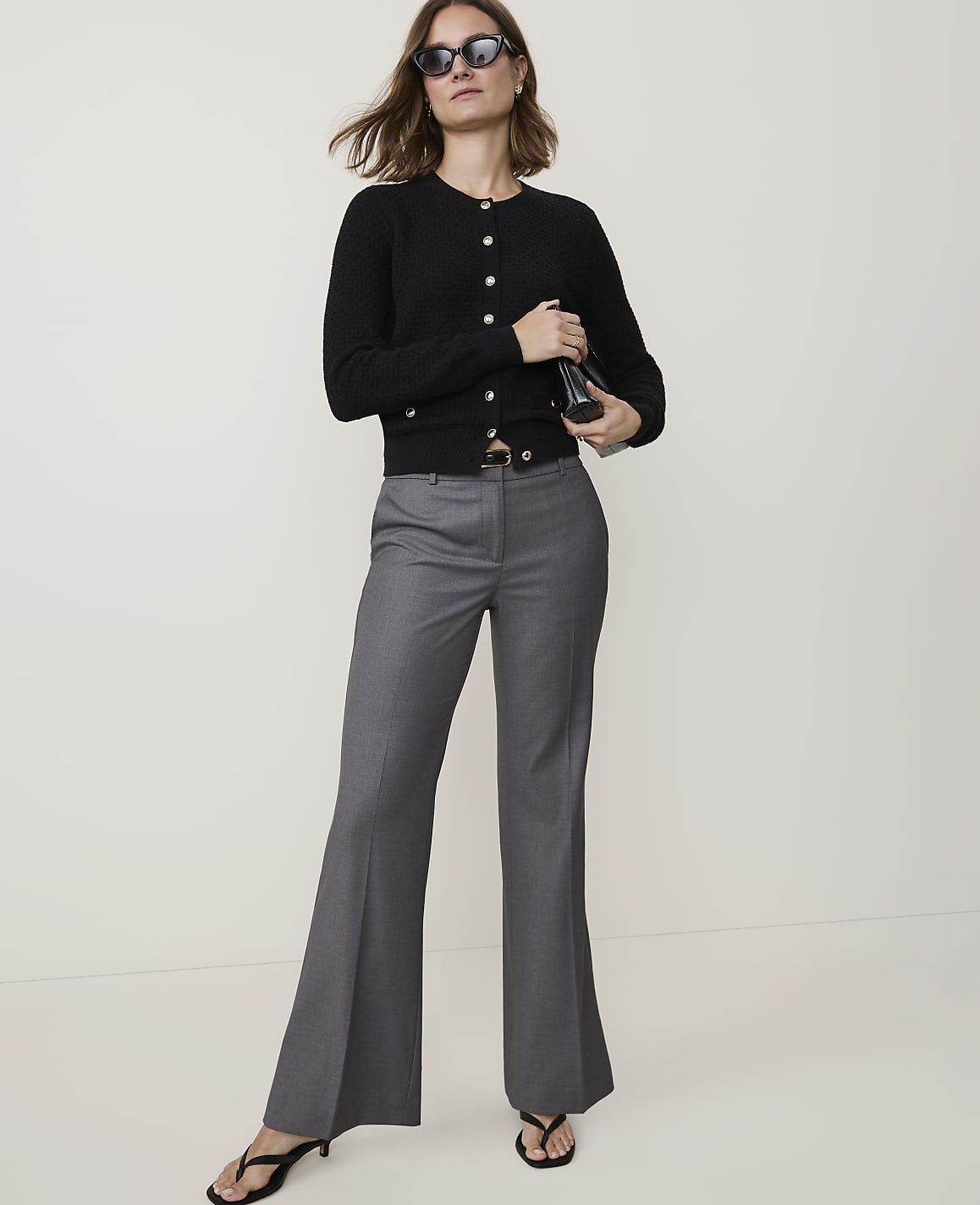 A woman in a black cardigan and gray flared pants stands against a plain, light-colored background.
