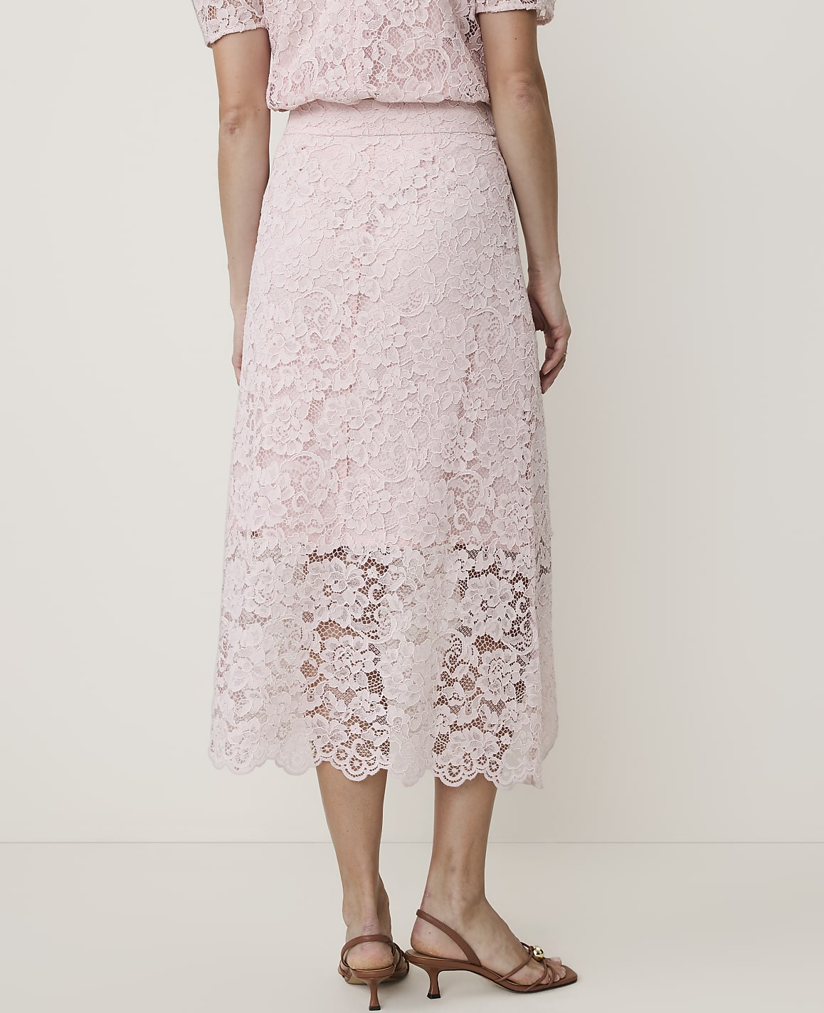 A person stands in a light pink lace skirt and top, with a plain white background.