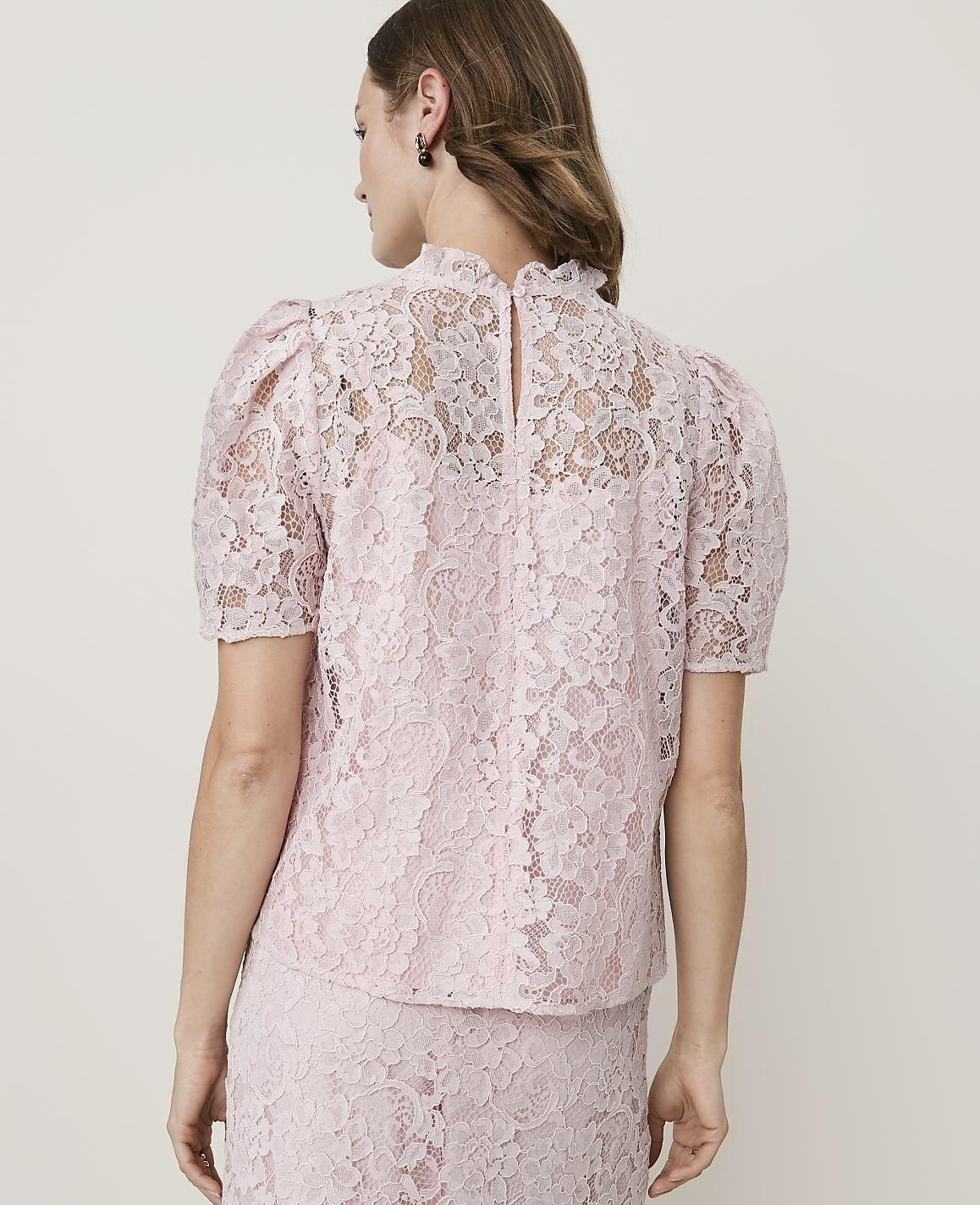 A woman in a pink lace top with puffed sleeves stands against a plain white background.