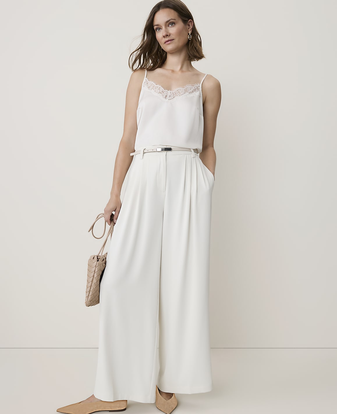 A woman in a white camisole and wide-leg pants stands against a plain, light background, holding a woven purse.