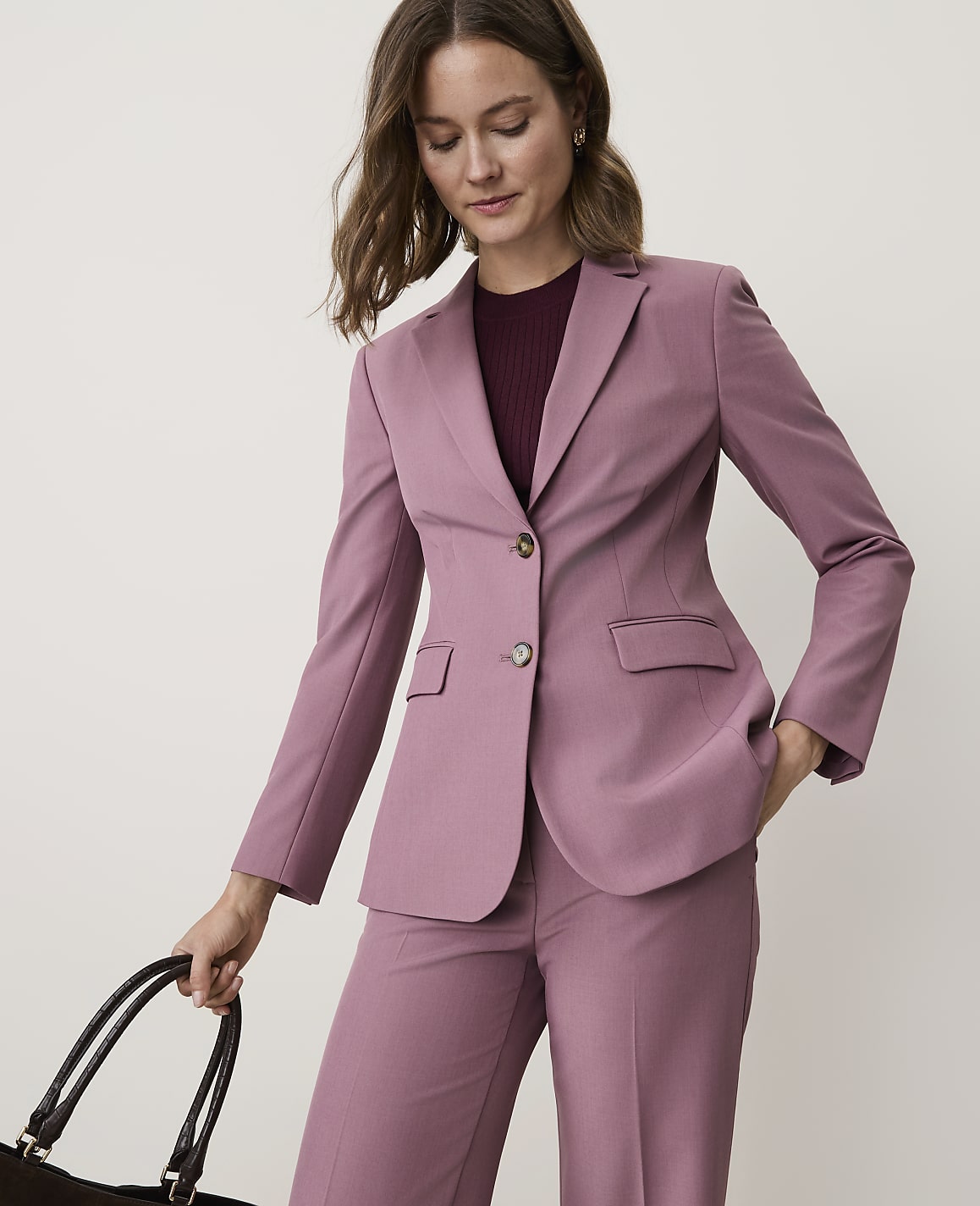 A woman in a mauve pantsuit with her hand in her pocket stands against a plain, light background, holding a dark tote bag.