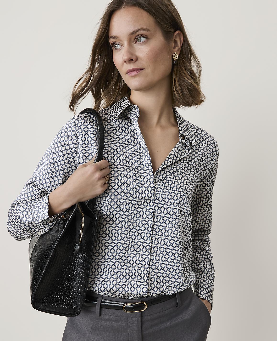 A woman in a patterned blouse and gray pants carries a black handbag against a plain background.