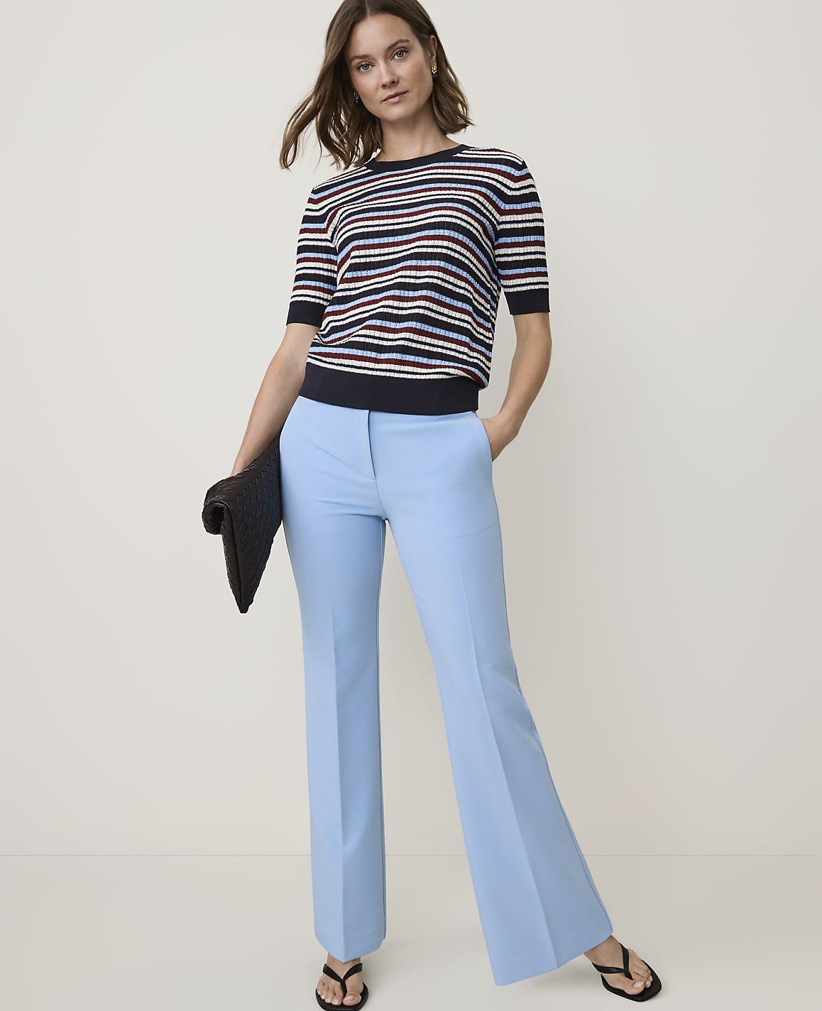 A woman in a striped sweater and light blue flared pants stands against a plain, light background, holding a dark clutch.