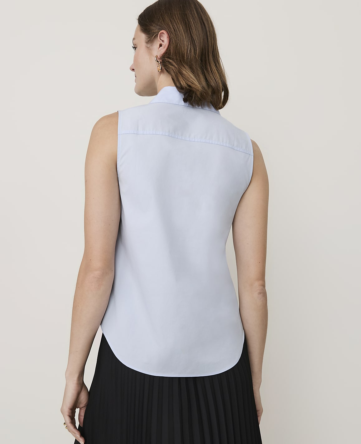 A woman in a light blue sleeveless collared shirt and a black pleated skirt stands against a plain white background.