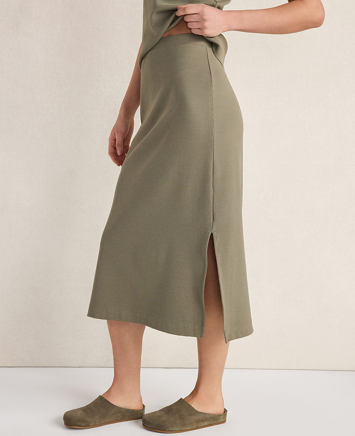 A person wearing a ribbed olive green midi skirt and clogs stands against a neutral, textured background.