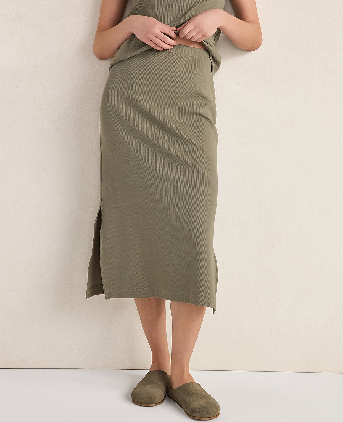 A person stands in a ribbed olive green midi skirt and matching top, wearing suede clogs, against a plain, light-colored wall.