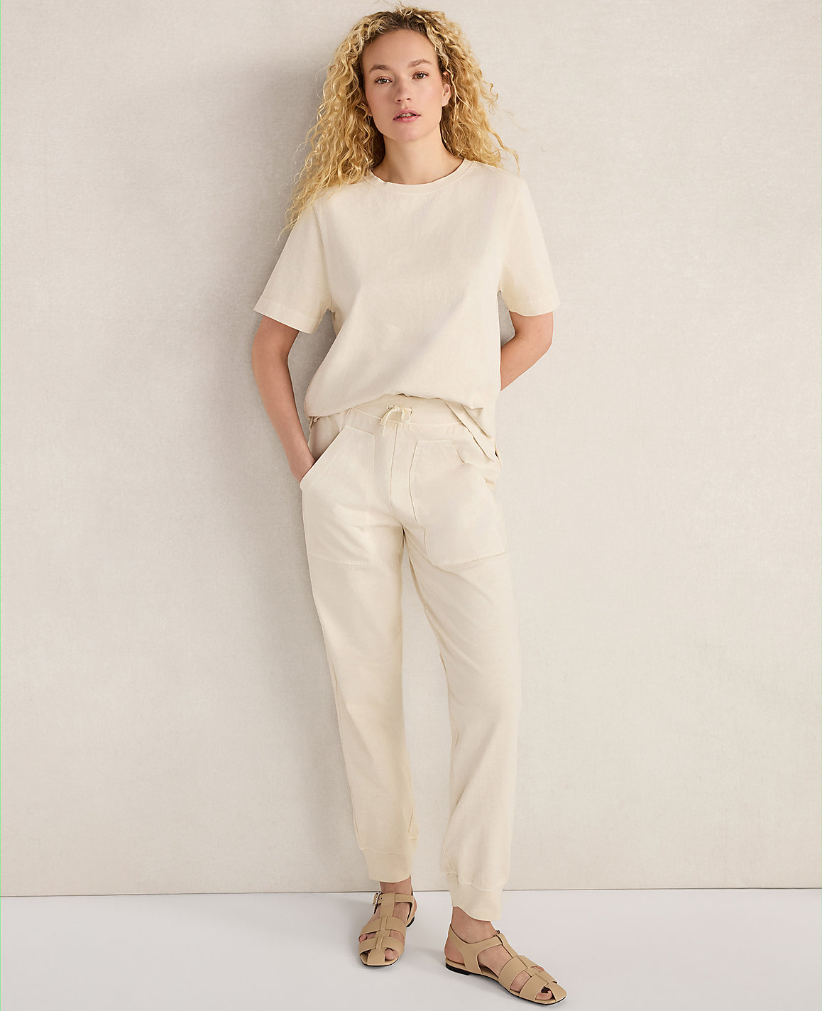 A woman in a cream-colored outfit stands against a textured, light-colored wall.