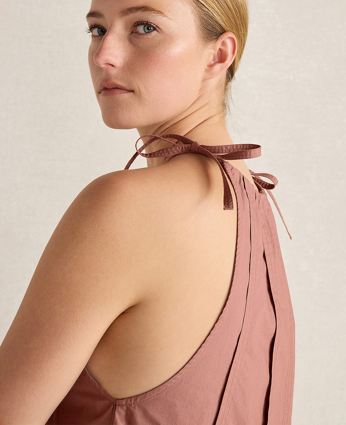 A woman with blonde hair tied in a bun wears a dusty rose halter top with ribbon ties, against a plain, light-colored background.