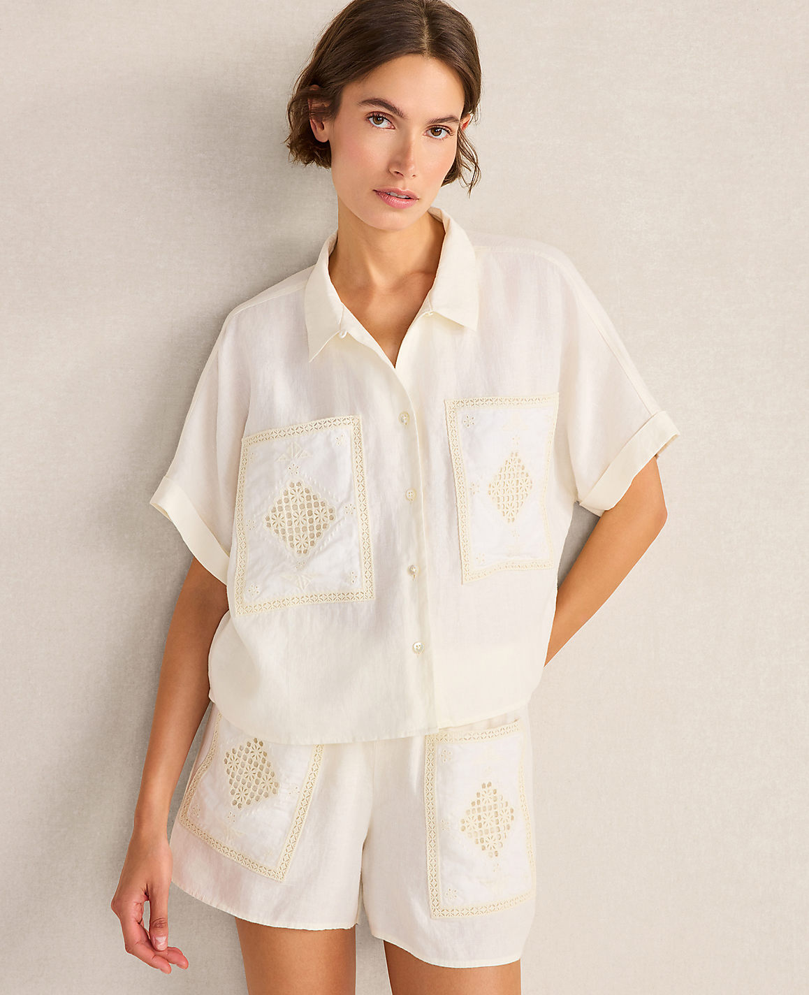 A woman in a white linen shirt and shorts with embroidered diamond patterns stands against a textured, light-colored wall.