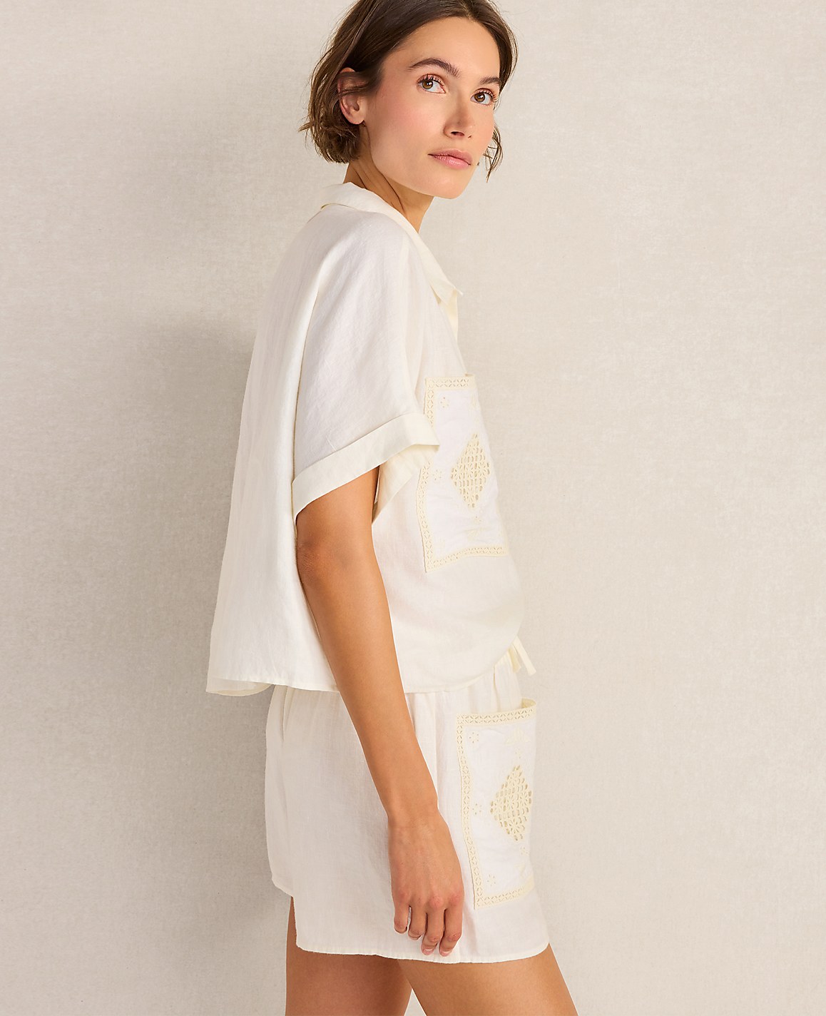 A woman in a white pajama set with embroidered details stands against a plain, light-colored background.