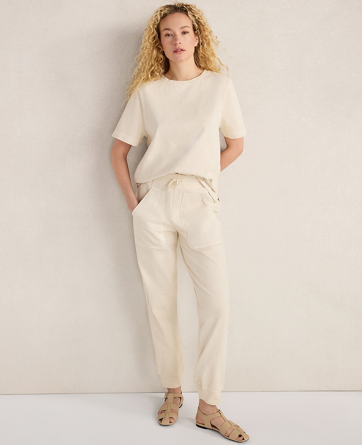 A woman with curly blonde hair stands in a cream-colored outfit against a textured off-white wall.