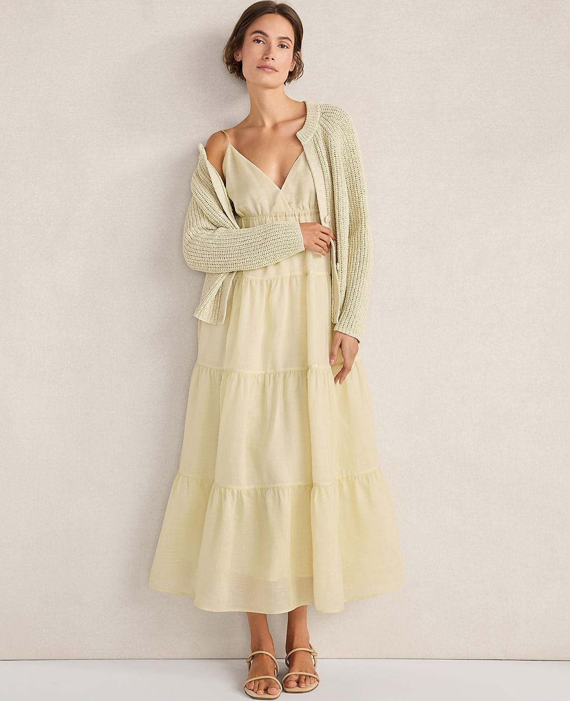 A woman in a light yellow tiered dress and a cream cardigan stands against a plain, light-colored wall.