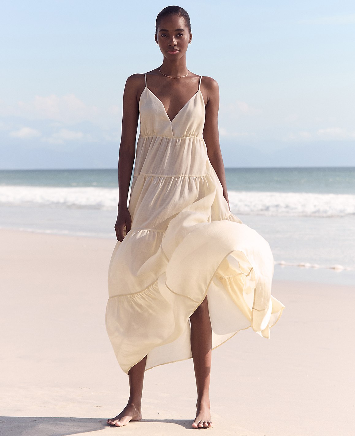 A woman in a flowing cream dress stands on a sandy beach with gentle waves and a clear sky in the background.