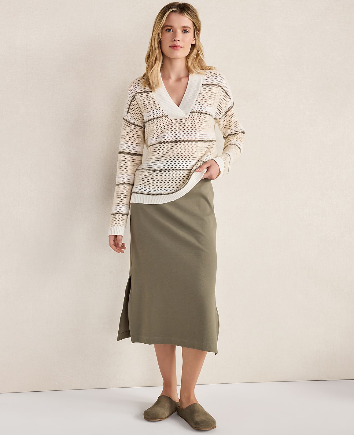 A woman in a striped sweater and olive skirt stands against a textured cream wall.