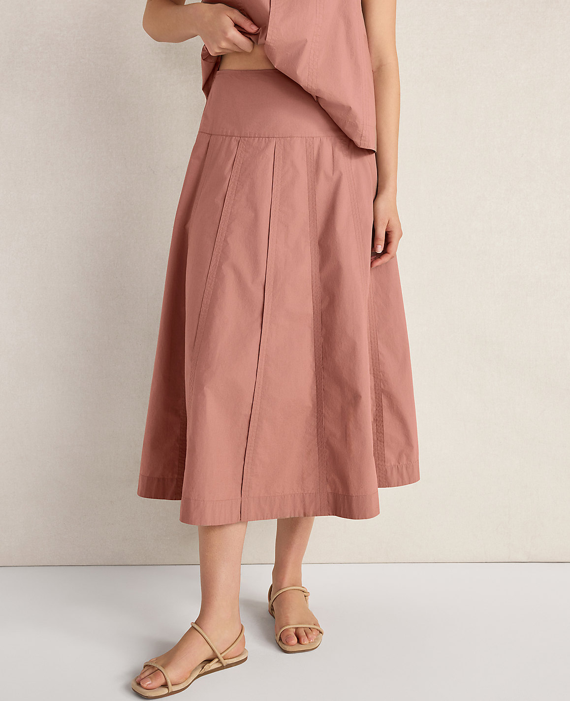 A person stands in a dusty rose pleated skirt and simple sandals against a textured off-white background.