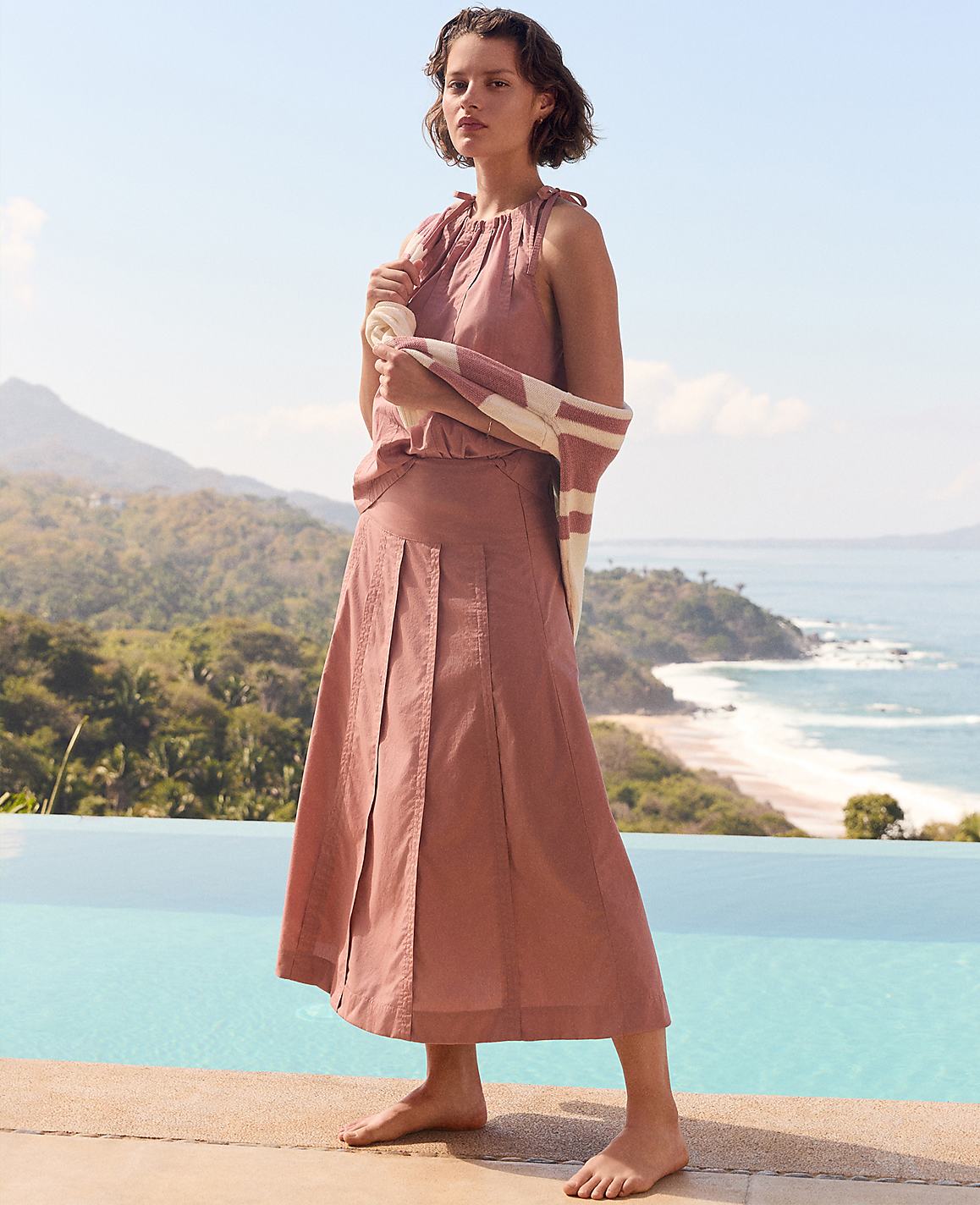 A woman in a pink dress stands by an infinity pool with a beach and lush green hills in the background.