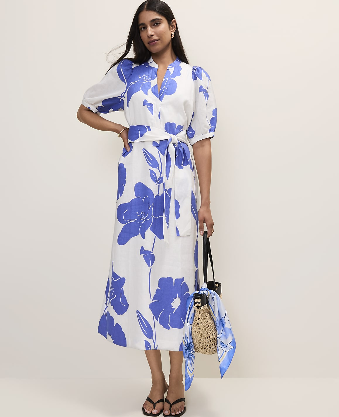 A woman in a white and blue floral dress stands in front of a plain, light-colored background, holding a straw bag with a scarf.