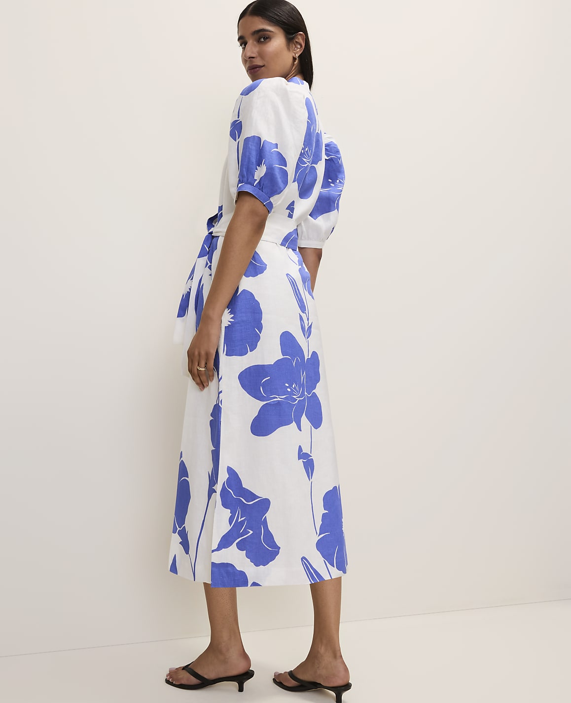 A woman in a white dress with large blue floral prints stands against a plain white background.