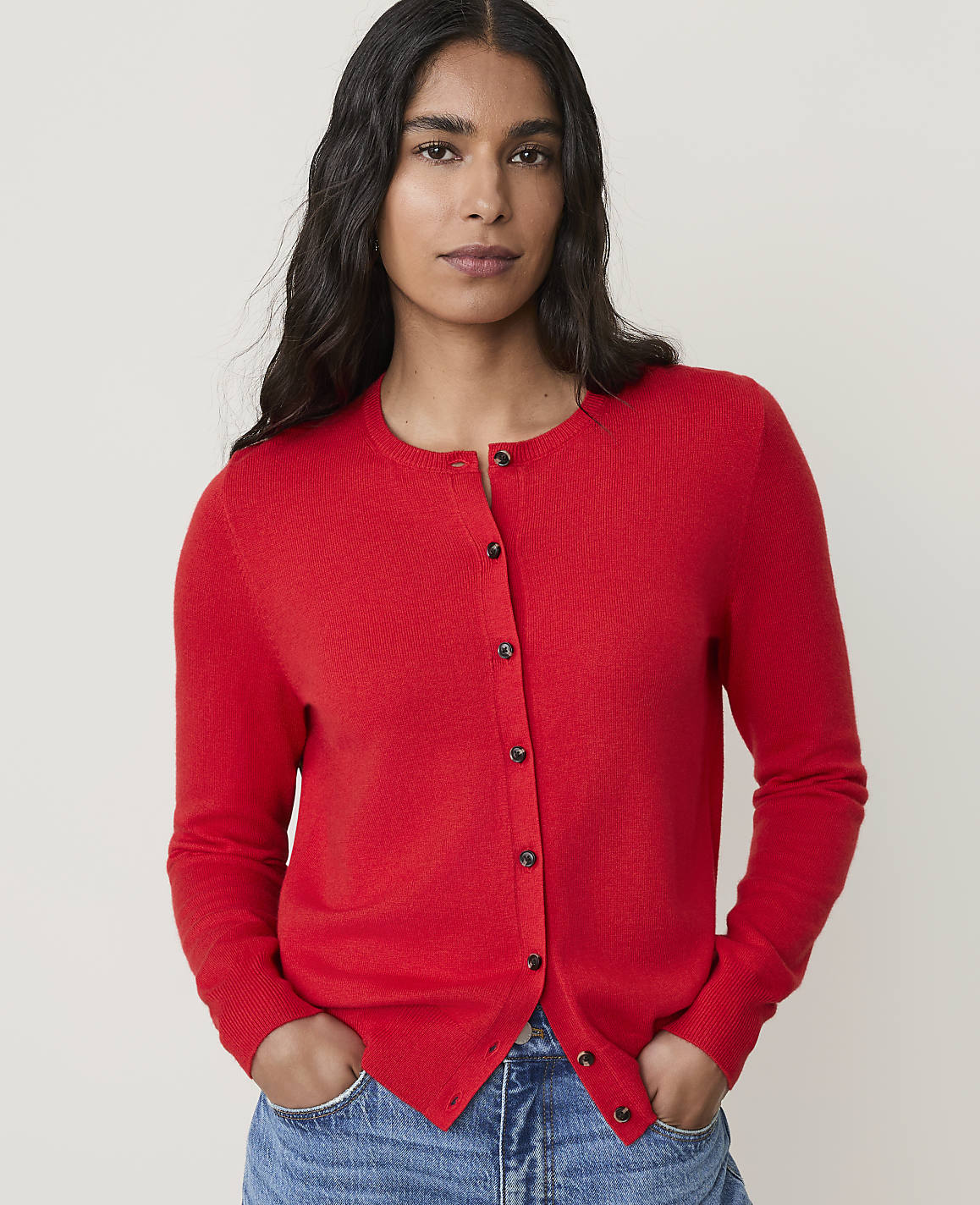 A woman in a red cardigan and blue jeans stands against a plain white background.
