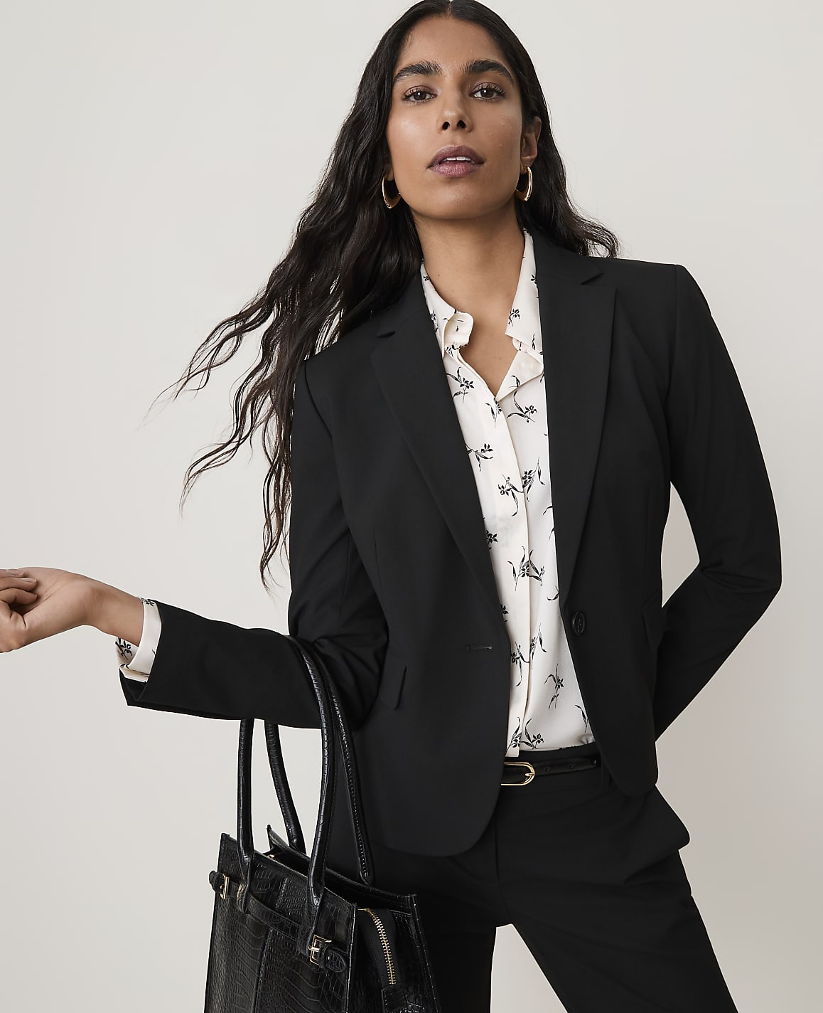A woman in a black blazer and patterned blouse holds a black handbag in front of a plain white background.