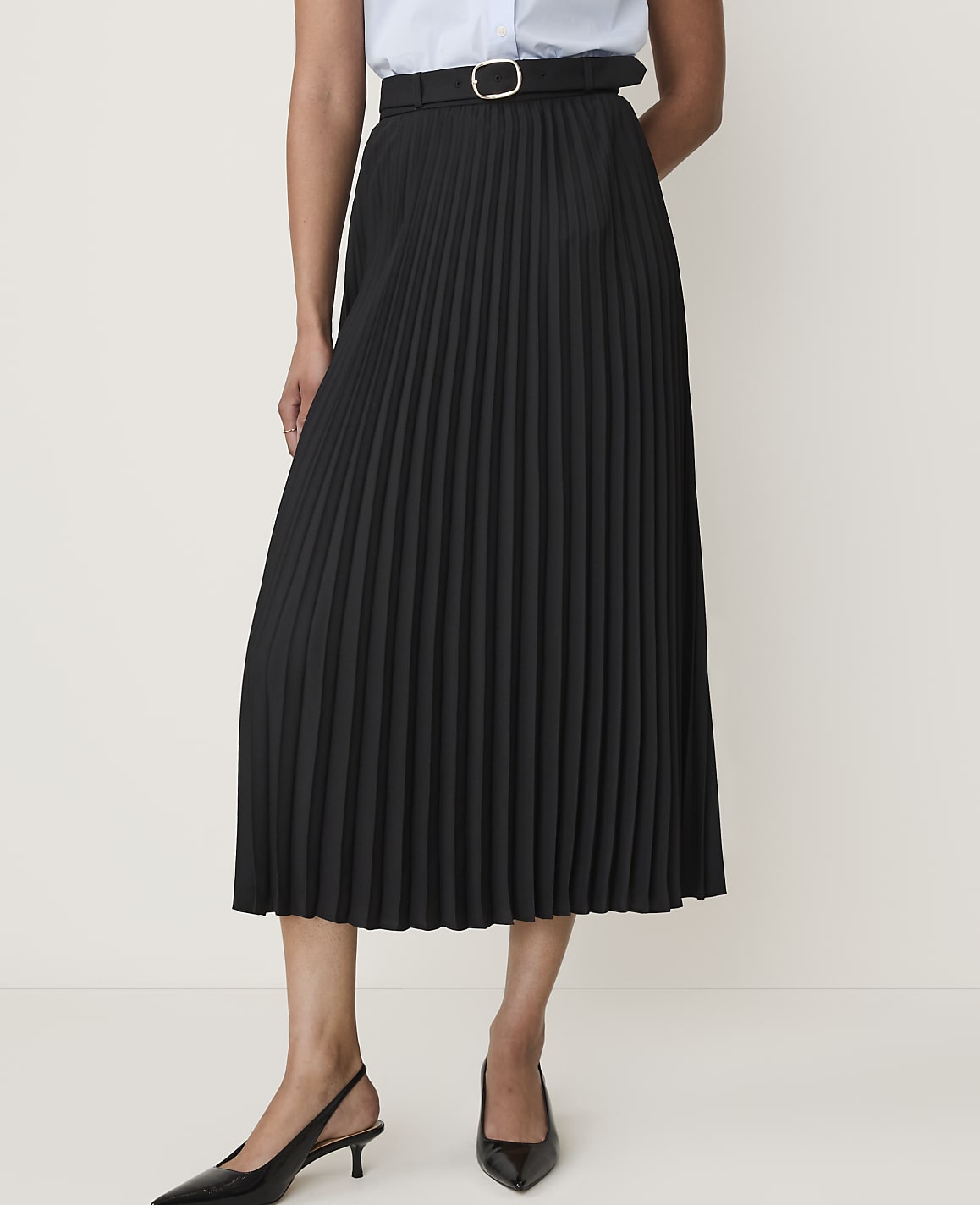 A person models a black pleated midi skirt with a belt and black slingback heels against a plain white background.