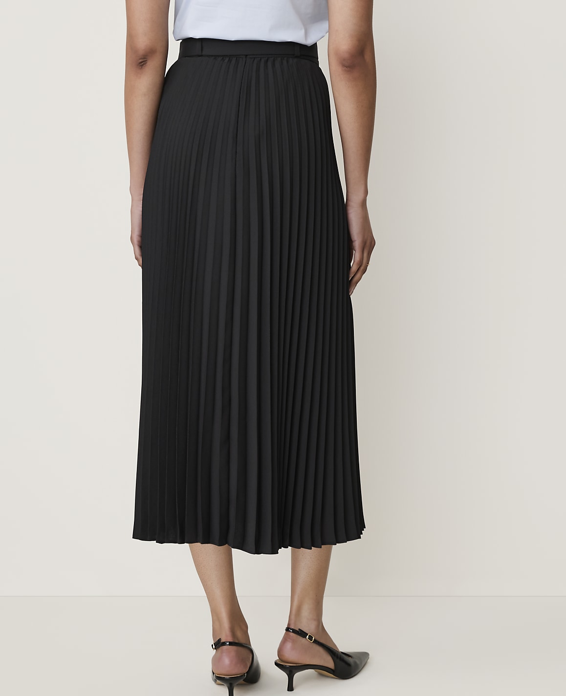 A person wearing a black pleated skirt and black heels stands against a plain, light-colored background.