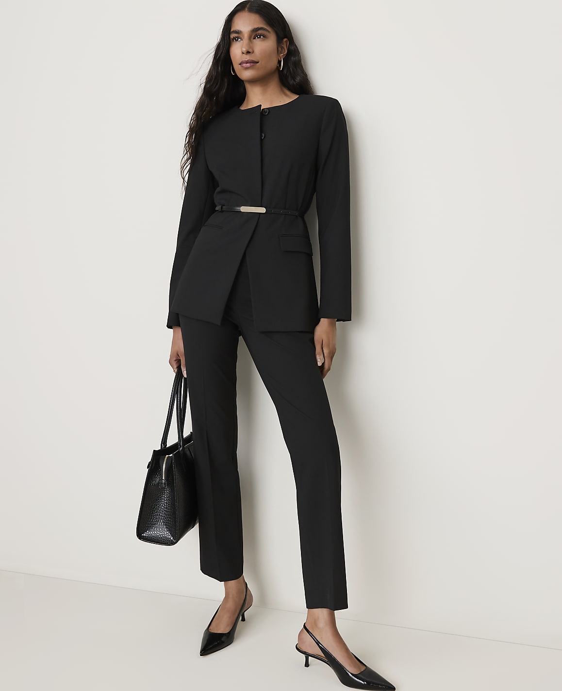 A woman in a black suit and heels stands with a handbag in front of a plain white wall.