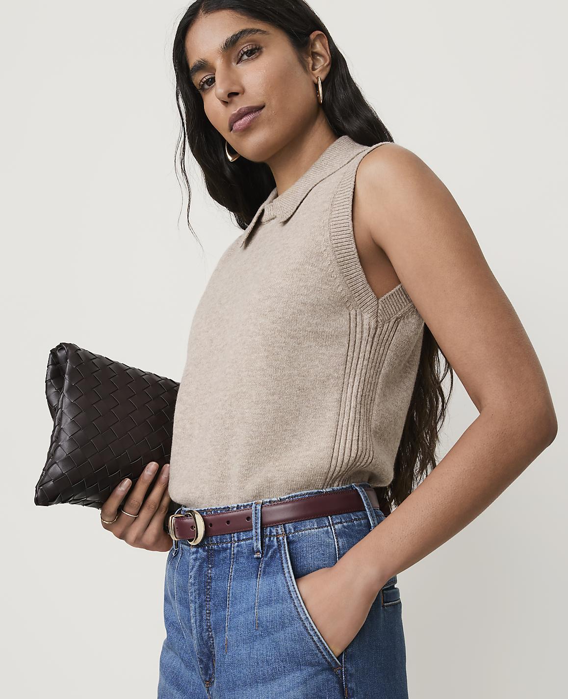 A woman in a beige collared vest and denim jeans holds a dark brown woven clutch against a plain background.