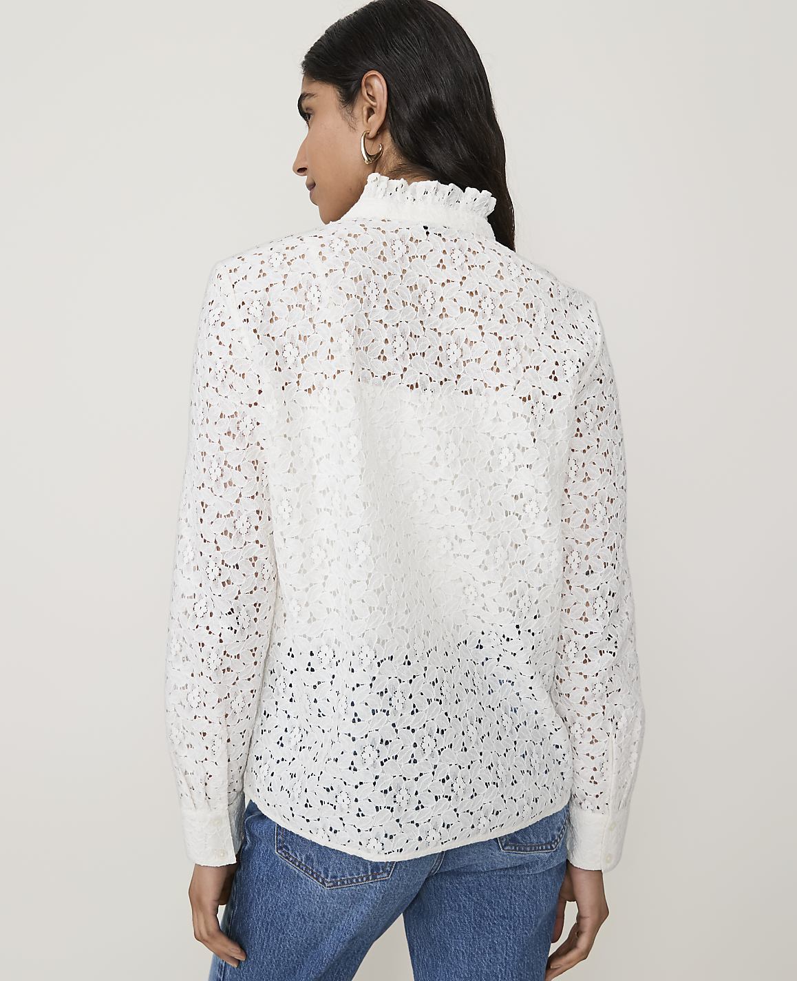 A woman in a white lace blouse and blue jeans stands against a plain white background.