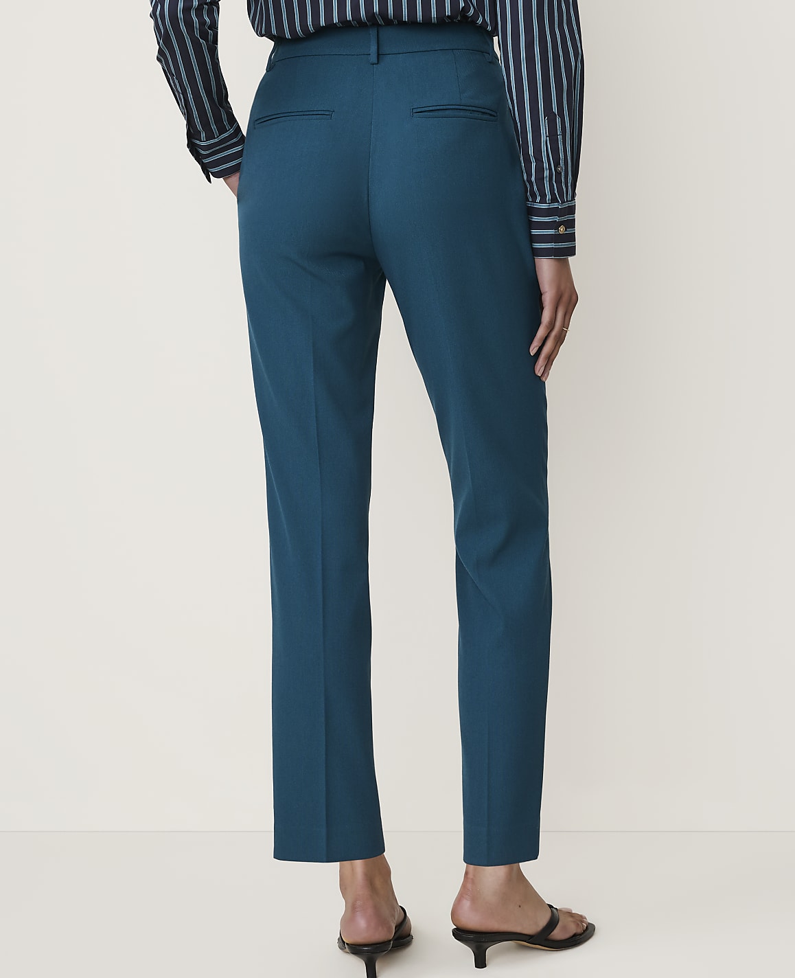 A person wearing teal trousers and a striped shirt stands against a plain white background.