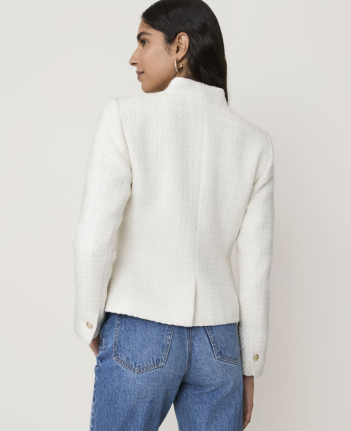 A woman in a white tweed jacket and blue jeans stands against a plain white background.