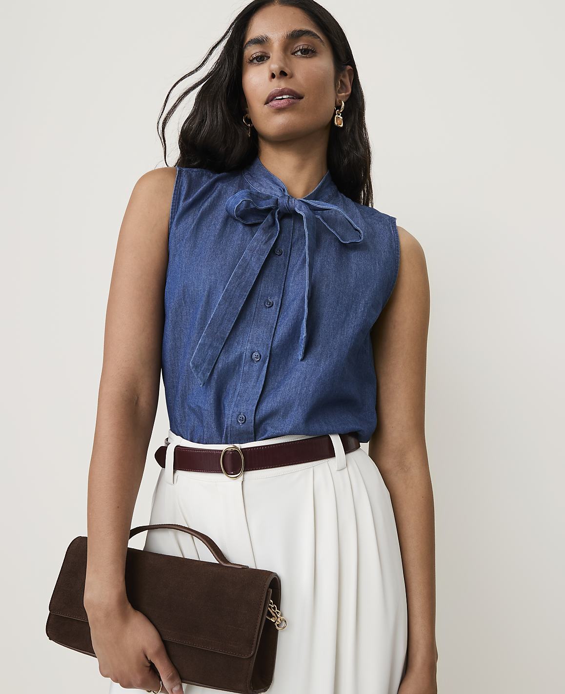 A woman in a denim bow-neck blouse and white pants holds a brown clutch in front of a plain background.