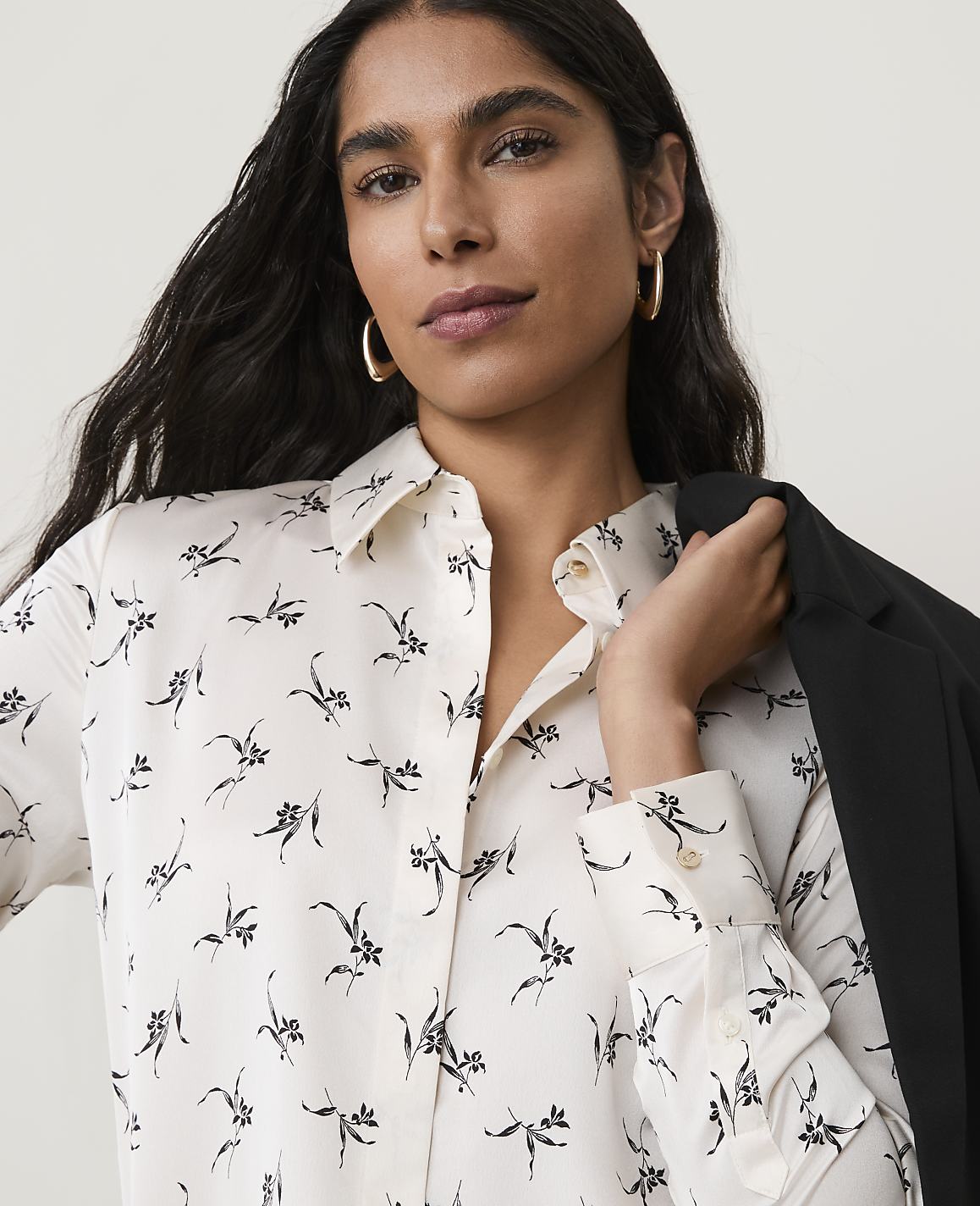 A woman with dark hair and hoop earrings wears a cream-colored, floral-patterned blouse and a black blazer draped over her shoulder, with a blurred background.