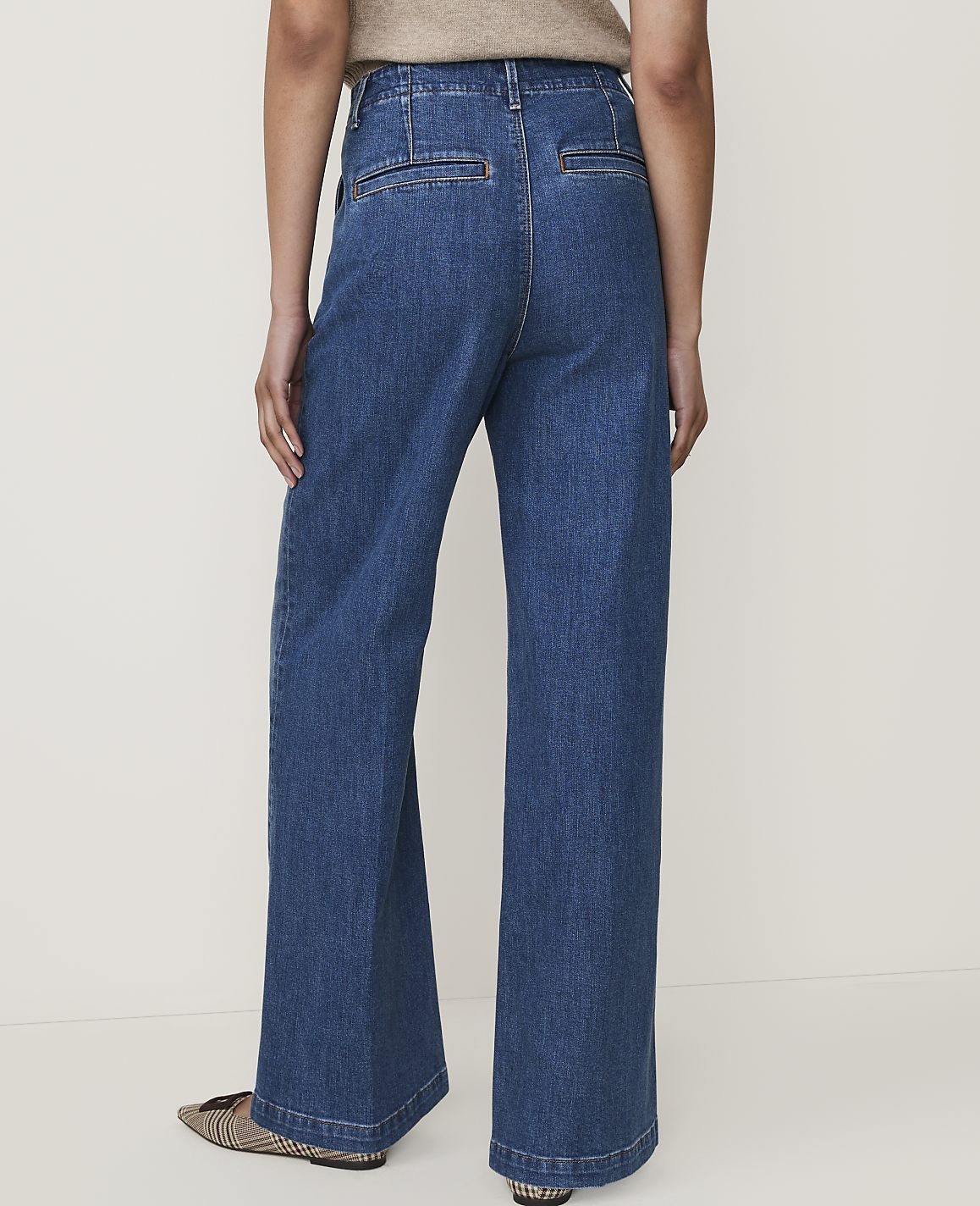 A person wearing wide-leg denim jeans and patterned flats stands against a plain, light-colored background.
