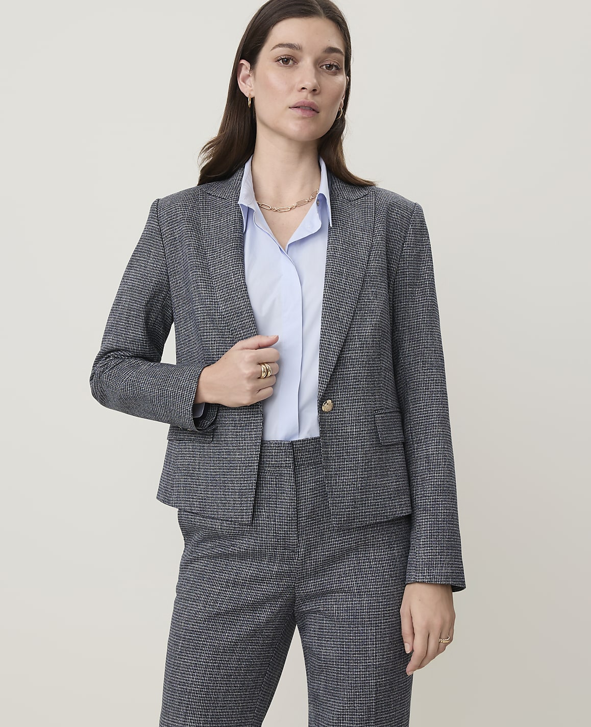 A woman in a grey patterned suit stands against a plain, light-colored background.