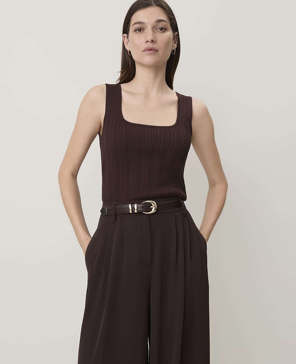 A woman in a brown ribbed tank top and wide-leg pants stands against a plain, light background.