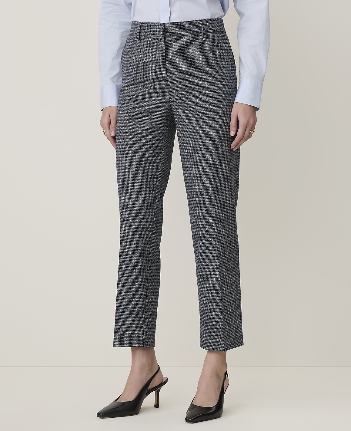 A person stands in a light gray room wearing a blue shirt and patterned gray pants with black heels.