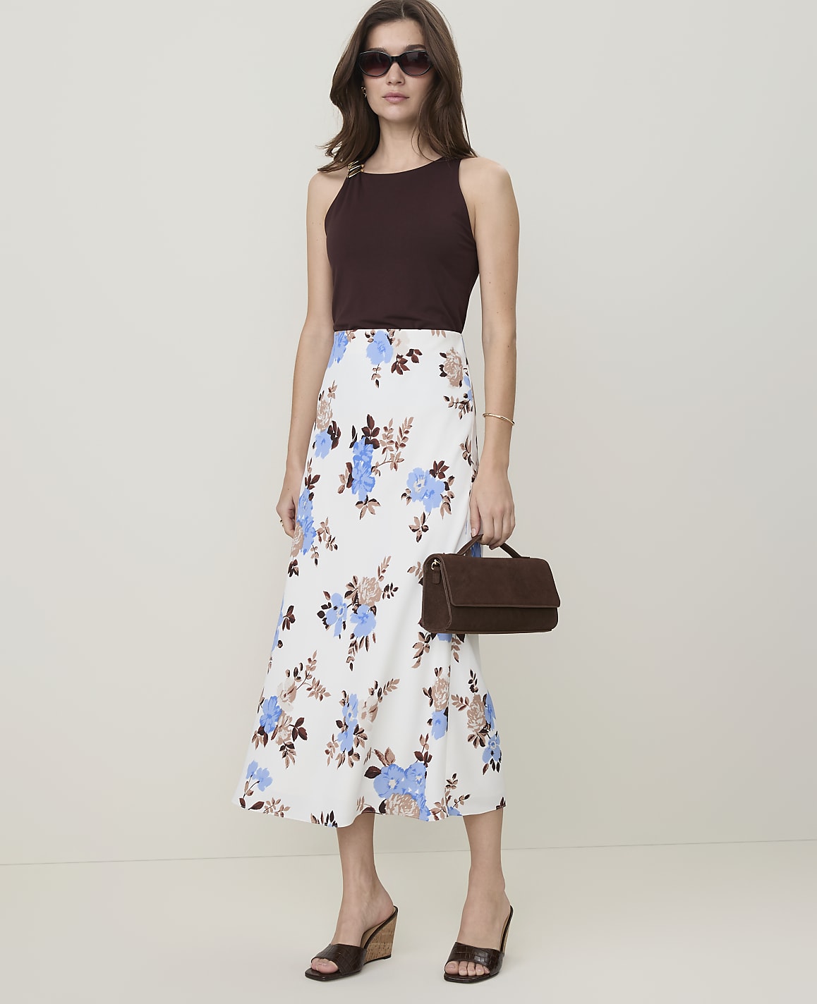 A woman in a floral skirt and dark top stands against a plain background, holding a brown handbag.