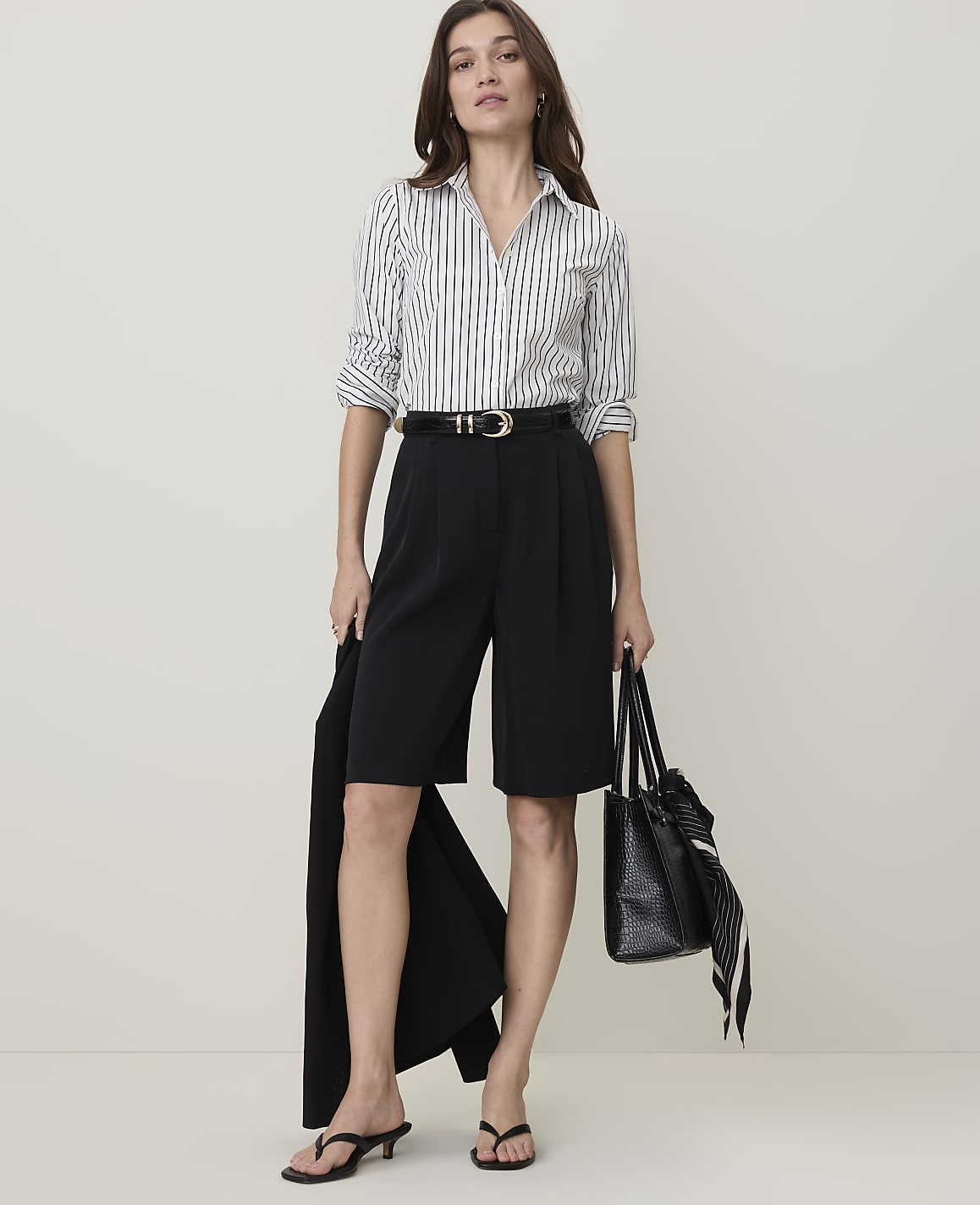 A woman in a striped shirt and black culottes stands against a plain, light background, holding a black blazer and a tote bag.