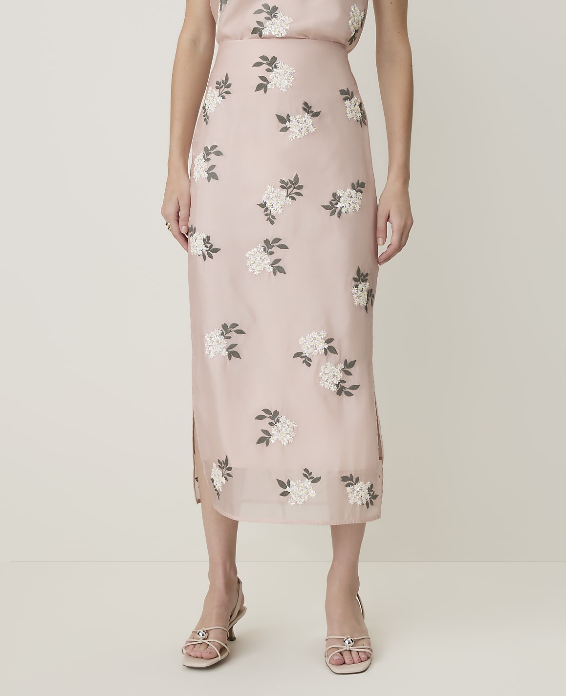 A person stands in front of a plain background, wearing a light pink floral embroidered midi skirt and light-colored sandals.