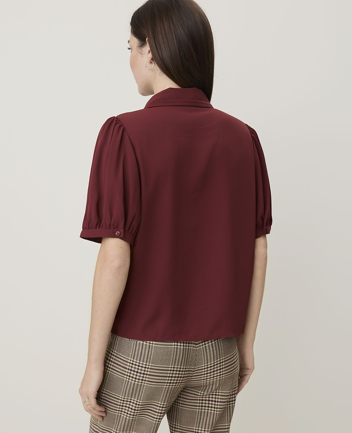 A woman in a maroon puff-sleeve blouse and plaid pants stands against a plain background.